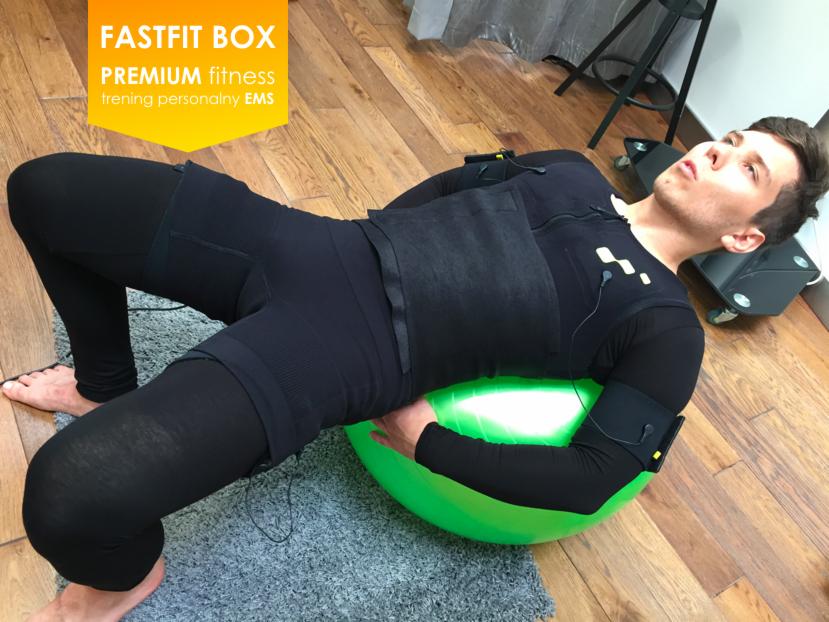 Mężczyzna w czarnym stroju EMS ćwiczy unoszenie bioder na zielonej piłce gimnastycznej, widoczne przewody i elektrody, w tle drewniana podłoga i sprzęt EMS.