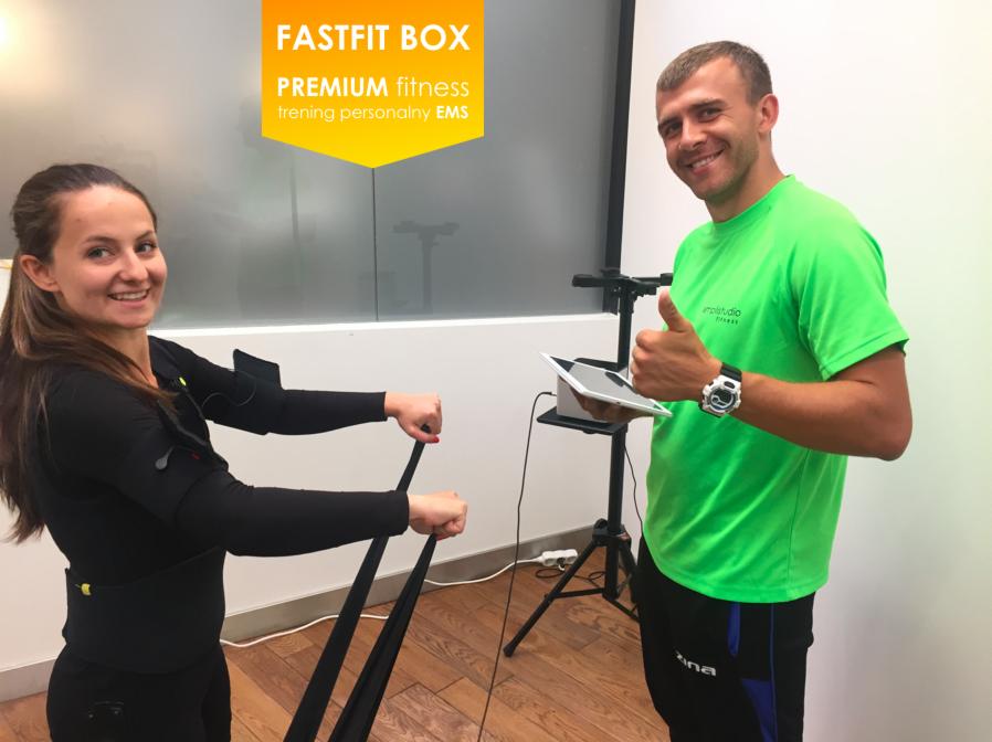 Kobieta w stroju EMS ćwiczy z gumami oporowymi pod okiem trenera z tabletem, który pokazuje kciuk w górę.