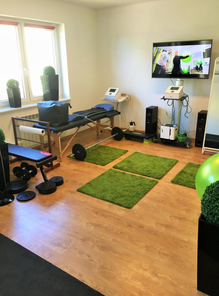 Wnętrze gabinetu z kozetką do masażu, sprzętem do ćwiczeń i telewizorem wyświetlającym ćwiczenia.