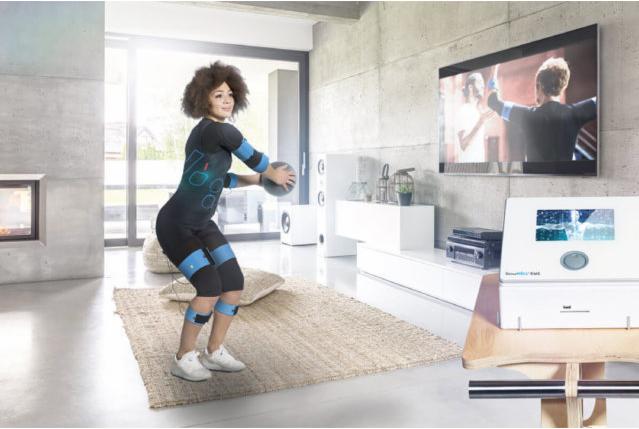 Kobieta w kombinezonie do elektrostymulacji mięśni ćwiczy z piłką w nowoczesnym wnętrzu, telewizor pokazuje sportowy mecz.