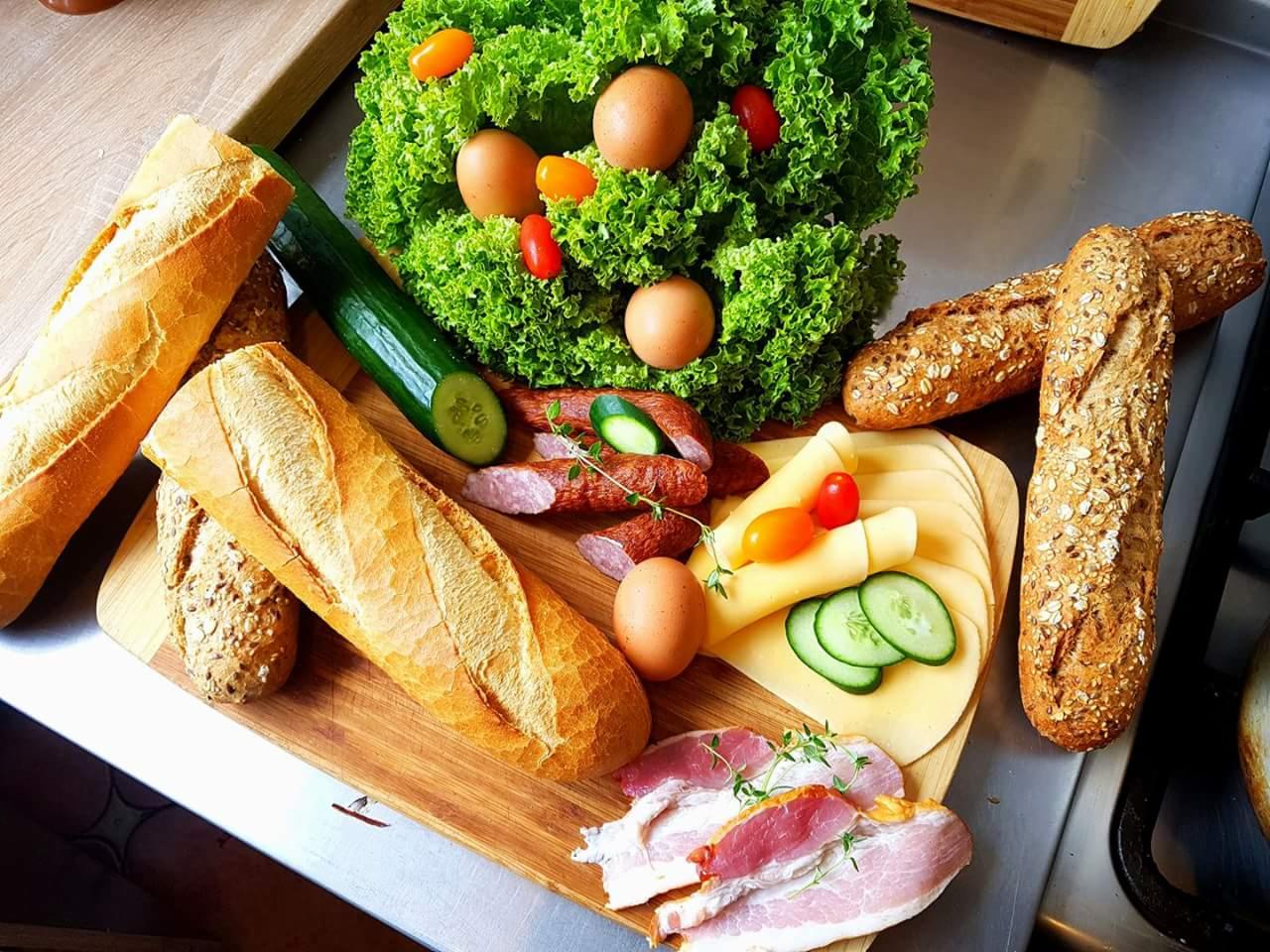 Drewniana deska z przygotowanymi składnikami do kanapek: bagietki, sałata, jajka, wędlina, ser, ogórek i pomidorki koktajlowe.