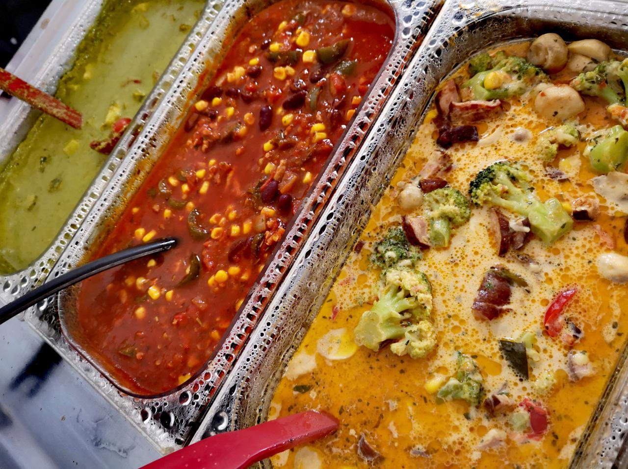 Trzy metalowe pojemniki GN wypełnione różnymi zupami: zielona kremowa, czerwona z kukurydzą i fasolą oraz żółta z brokułami i grzybami, widoczne łyżki do nakładania.