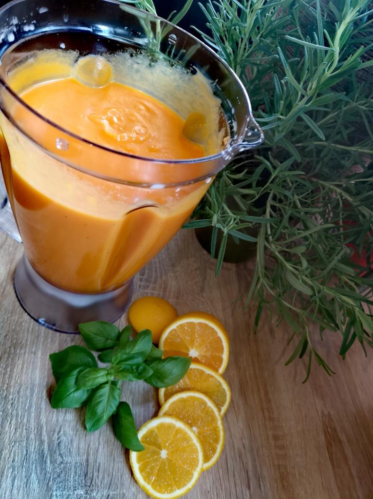 Kremowa pomarańczowa zupa w blenderze, otoczona plasterkami pomarańczy, cytryną i świeżą bazylią na drewnianym blacie, z gałązką rozmarynu w tle.