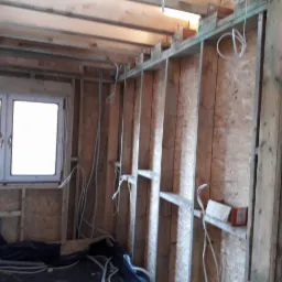 Wnętrze w trakcie budowy z widoczną drewnianą konstrukcją ścian, oknem z białymi ramami i przewodami elektrycznymi wystającymi ze ścian. W tle widoczne elementy instalacji pod sufitem.