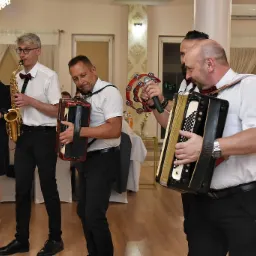 Muzycy grający na saksofonie, akordeonach i tamburynie podczas imprezy w sali z parkietem i żyrandolem.