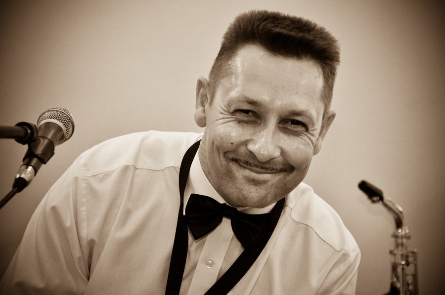 Portret uśmiechniętego mężczyzny z wąsami w białej koszuli i muszce, z mikrofonem i saksofonem w tle, utrzymany w sepii.