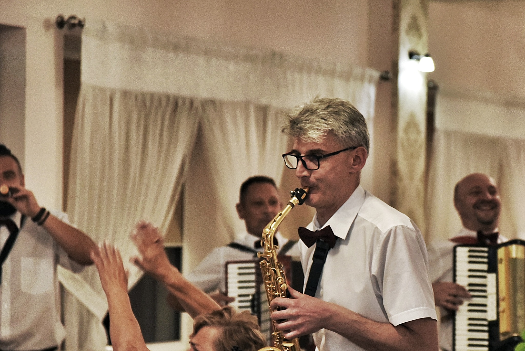 Saksofonista w okularach i muszce gra na saksofonie podczas wesela, za nim akordeonista i osoba z mikrofonem, w tle białe zasłony i podniesione ręce gości.