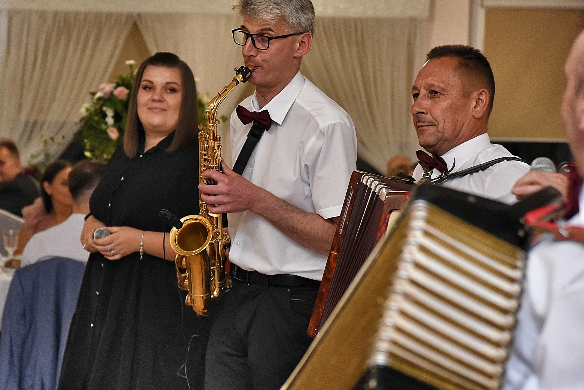 Saksofonista w okularach i muszce gra podczas wesela, obok wokalistka z mikrofonem i akordeonista, w tle dekoracja kwiatowa i goście.