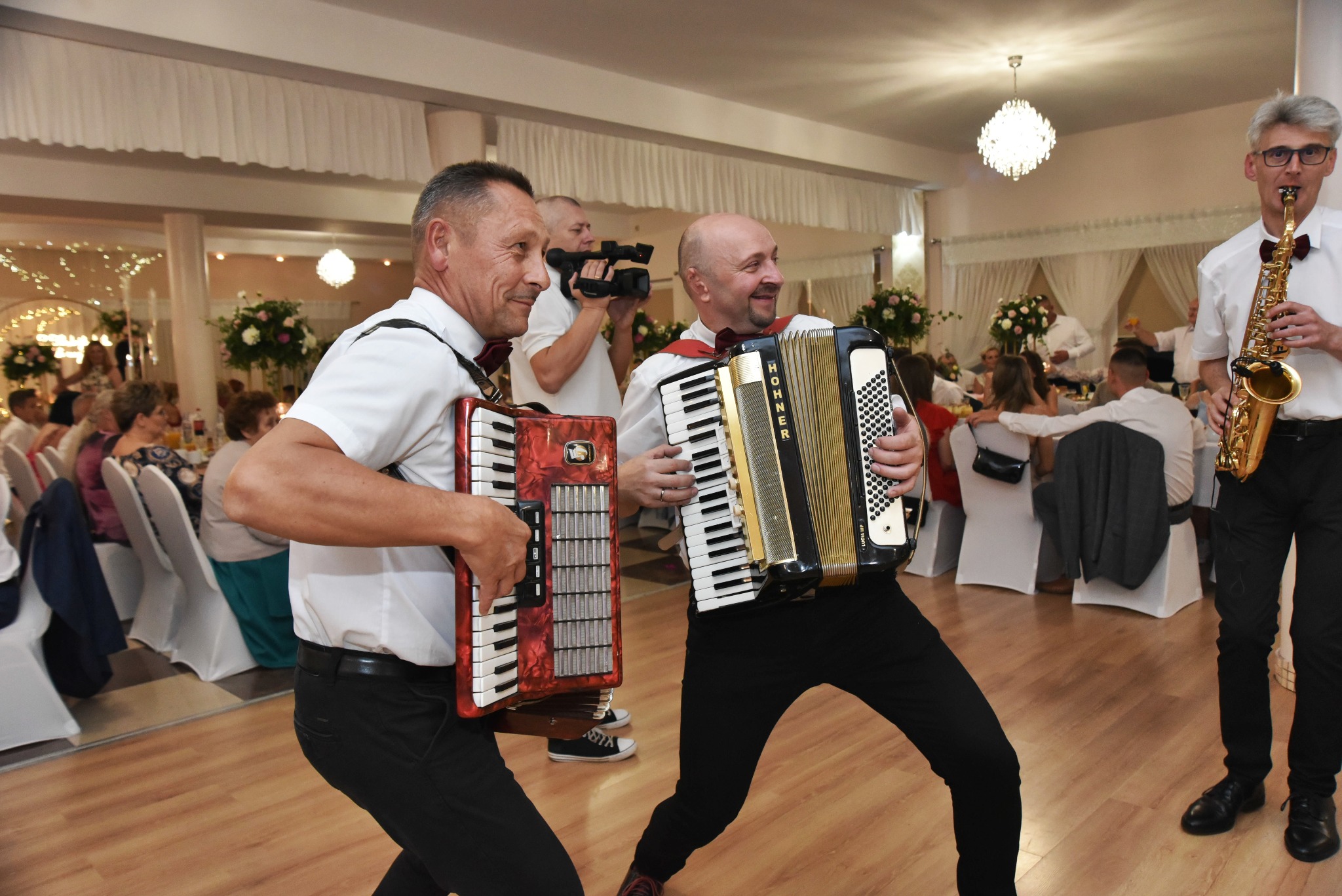 Muzycy grający na akordeonach i saksofonie podczas wesela w Brzeźniu, w tle widoczni goście i kamerzysta.