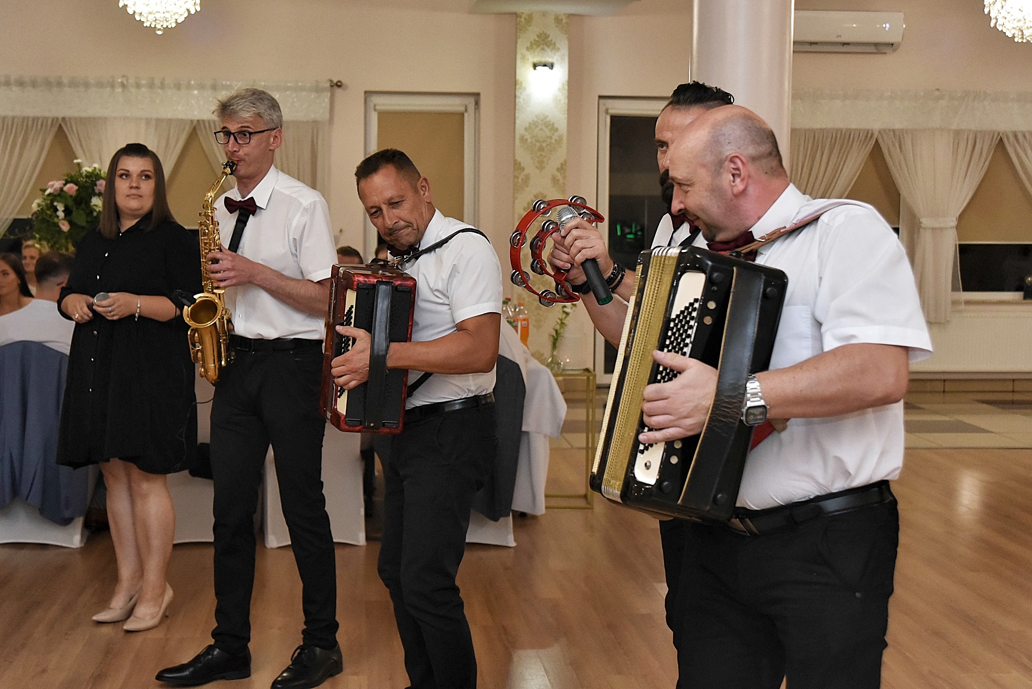 Muzycy grający na saksofonie, akordeonach i tamburynie podczas imprezy w sali z parkietem i żyrandolem.