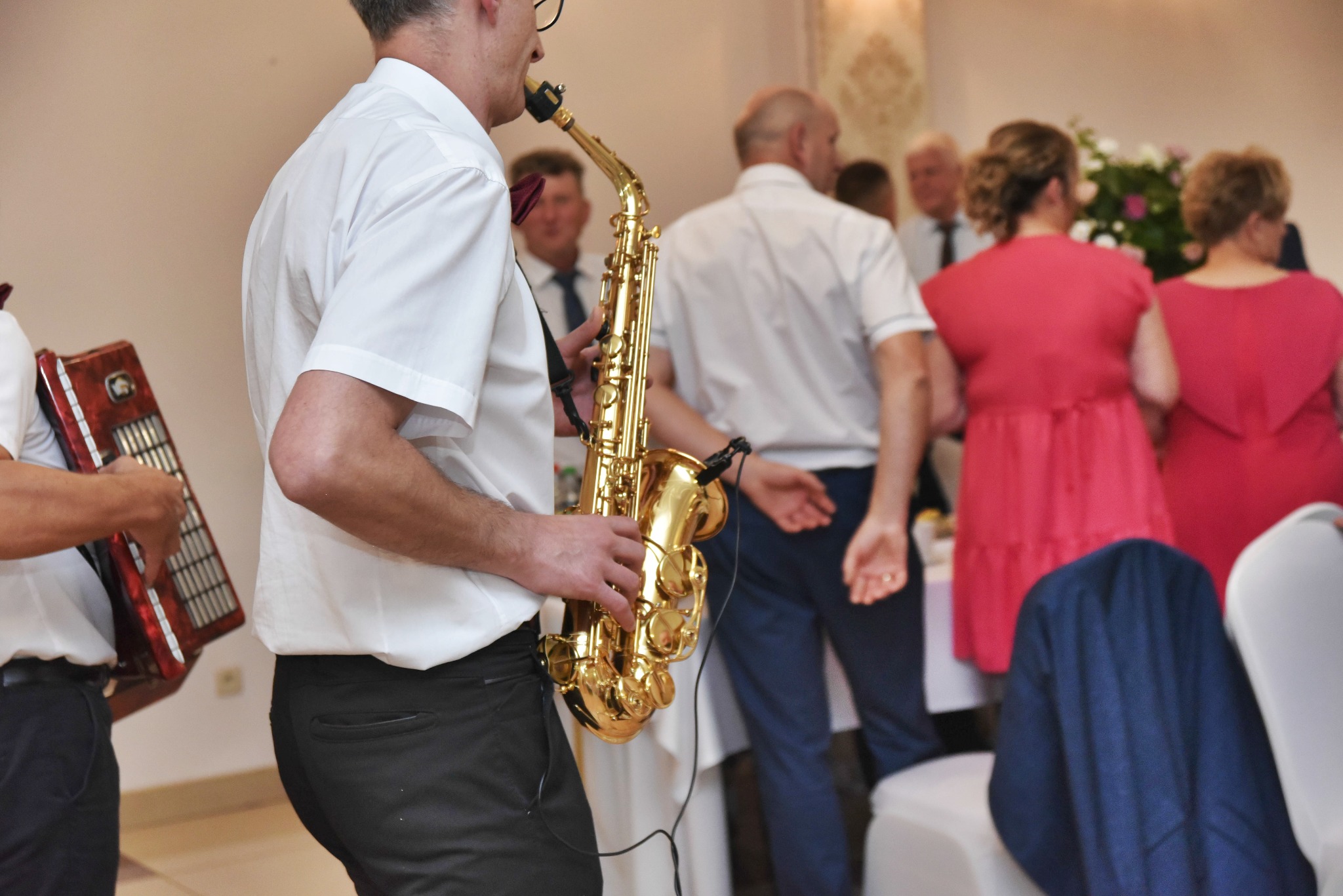 Saksofonista gra na złotym saksofonie podczas weselnej biesiady, w tle akordeonista i goście w strojach wieczorowych.