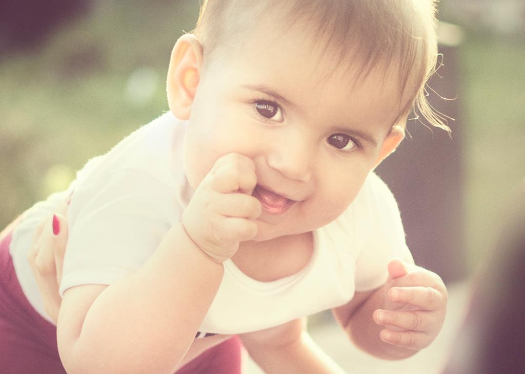 Uśmiechnięty niemowlak w białym body, z palcem w ustach, trzymany przez osobę z pomalowanymi na czerwono paznokciami, na rozmytym zielonym tle.