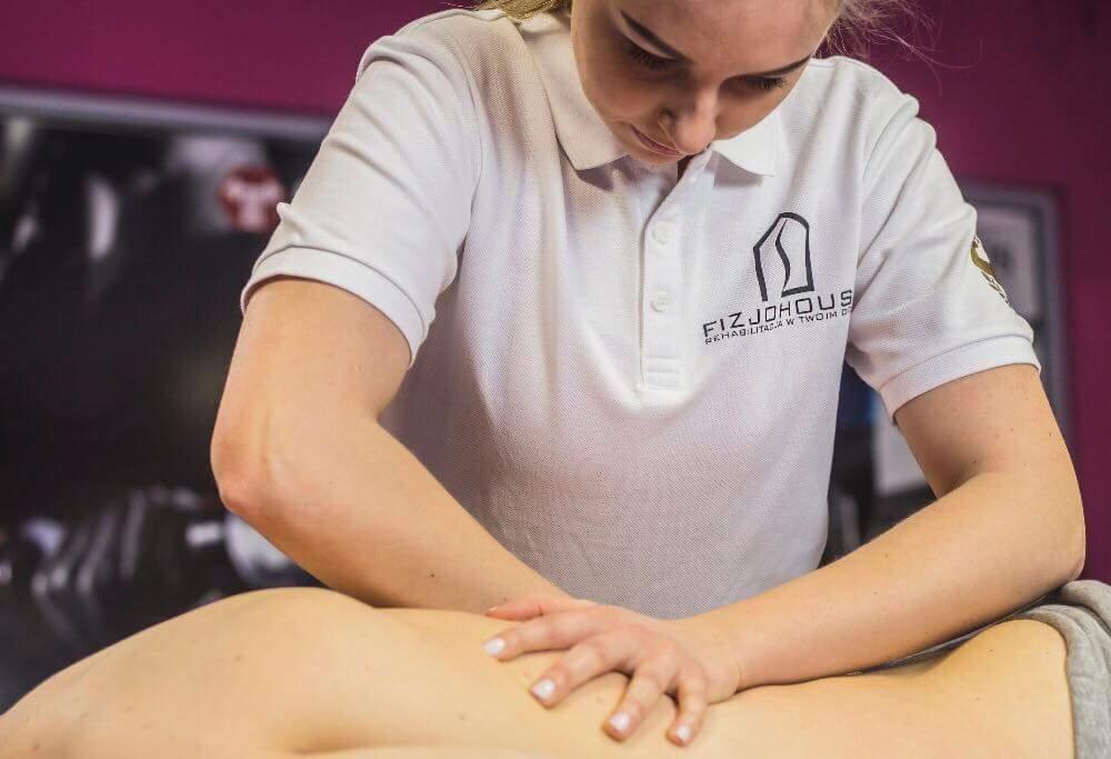 Kobieta w białej koszulce polo z logo firmy, wykonuje masaż pleców pacjenta, skupiona na precyzyjnych ruchach dłońmi.