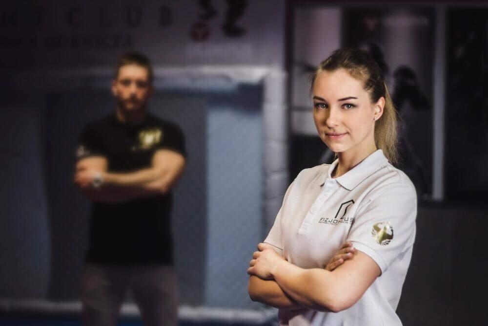 Portret uśmiechniętej kobiety w białej koszulce polo z logo firmy 'FIZJO PLUS', z mężczyzną w tle, oboje ze skrzyżowanymi ramionami w sali ćwiczeń.