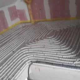 Białe rury układane spiralnie na metalowej siatce, przygotowane do zalania betonem, w trakcie instalacji systemu ogrzewania podłogowego w surowym wnętrzu poddasza z drewnianą konstrukcją dachu.