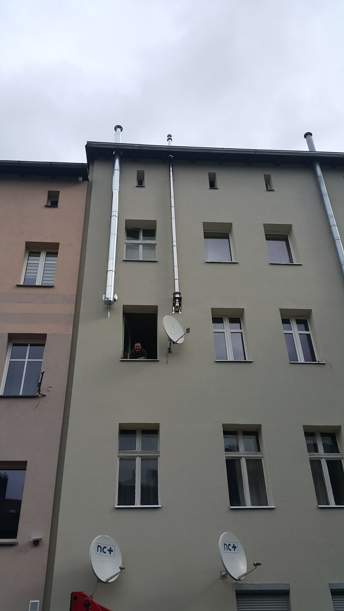 Zewnętrzny montaż pionowych, metalowych rur spalinowych na ścianie budynku wielorodzinnego, widoczny pracownik w oknie.