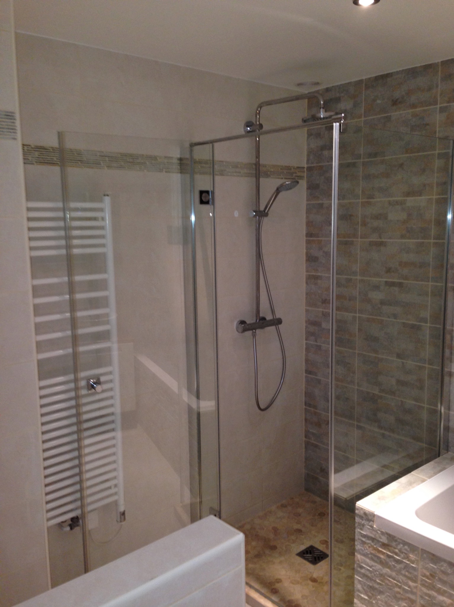 Nowoczesna kabina prysznicowa ze szklanymi drzwiami, grzejnik drabinkowy i płytki imitujące kamień w wykończonej łazience. Widoczny odpływ liniowy.