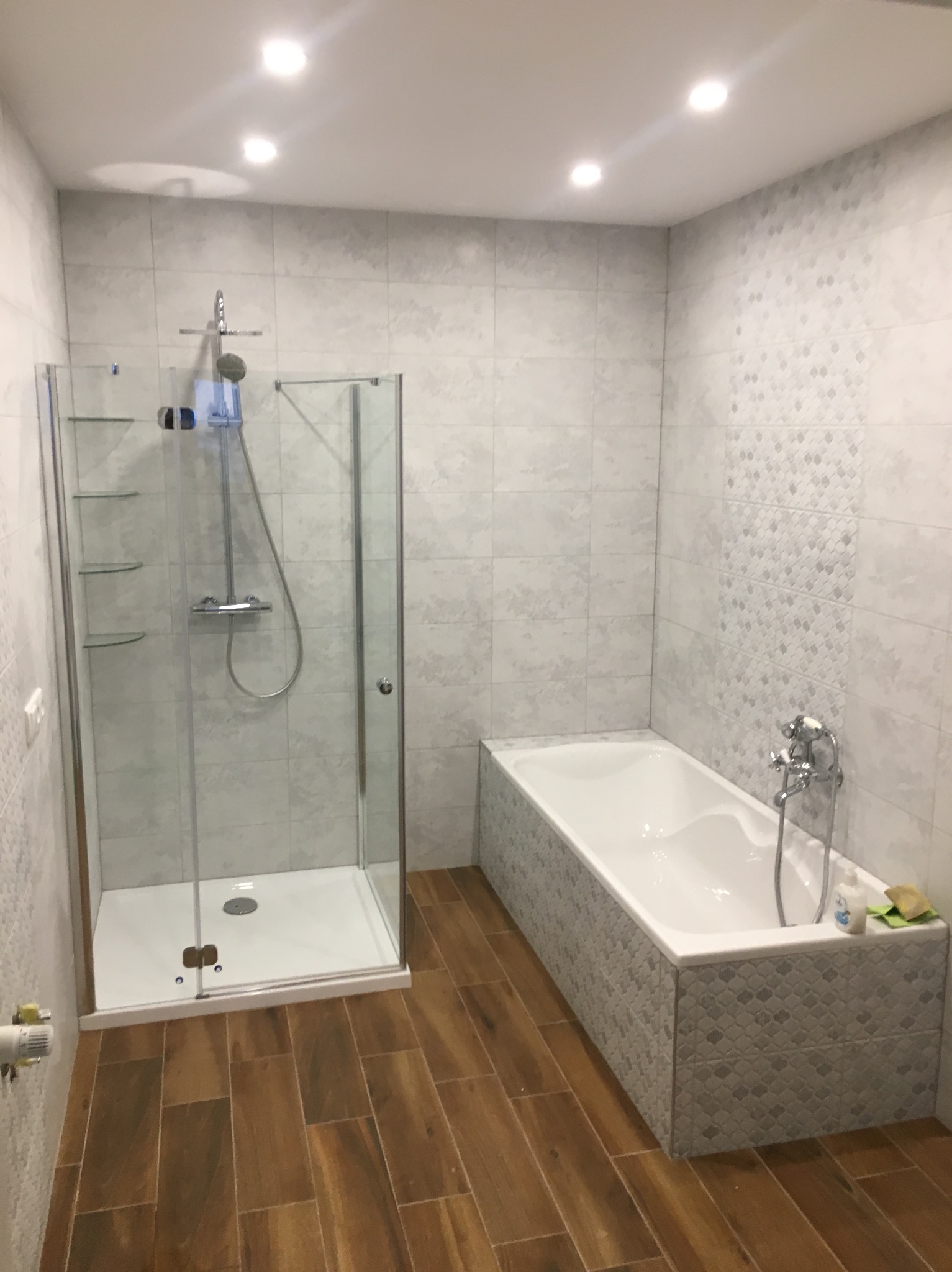 Nowoczesna łazienka po remoncie: kabina prysznicowa ze szklanymi półkami, wanna obudowana mozaiką i drewnopodobna podłoga. Jasne płytki na ścianach.