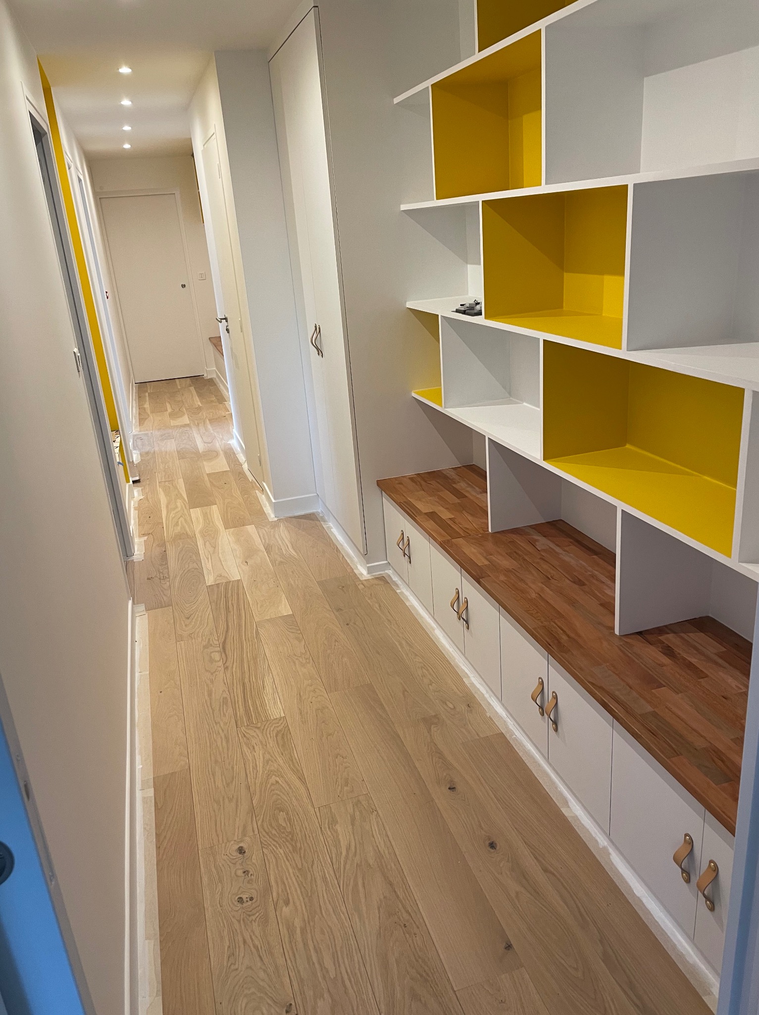Długi korytarz z drewnianą podłogą i zabudową: białe szafki z drewnianym blatem i regał z żółtymi akcentami. Oświetlenie punktowe w suficie.