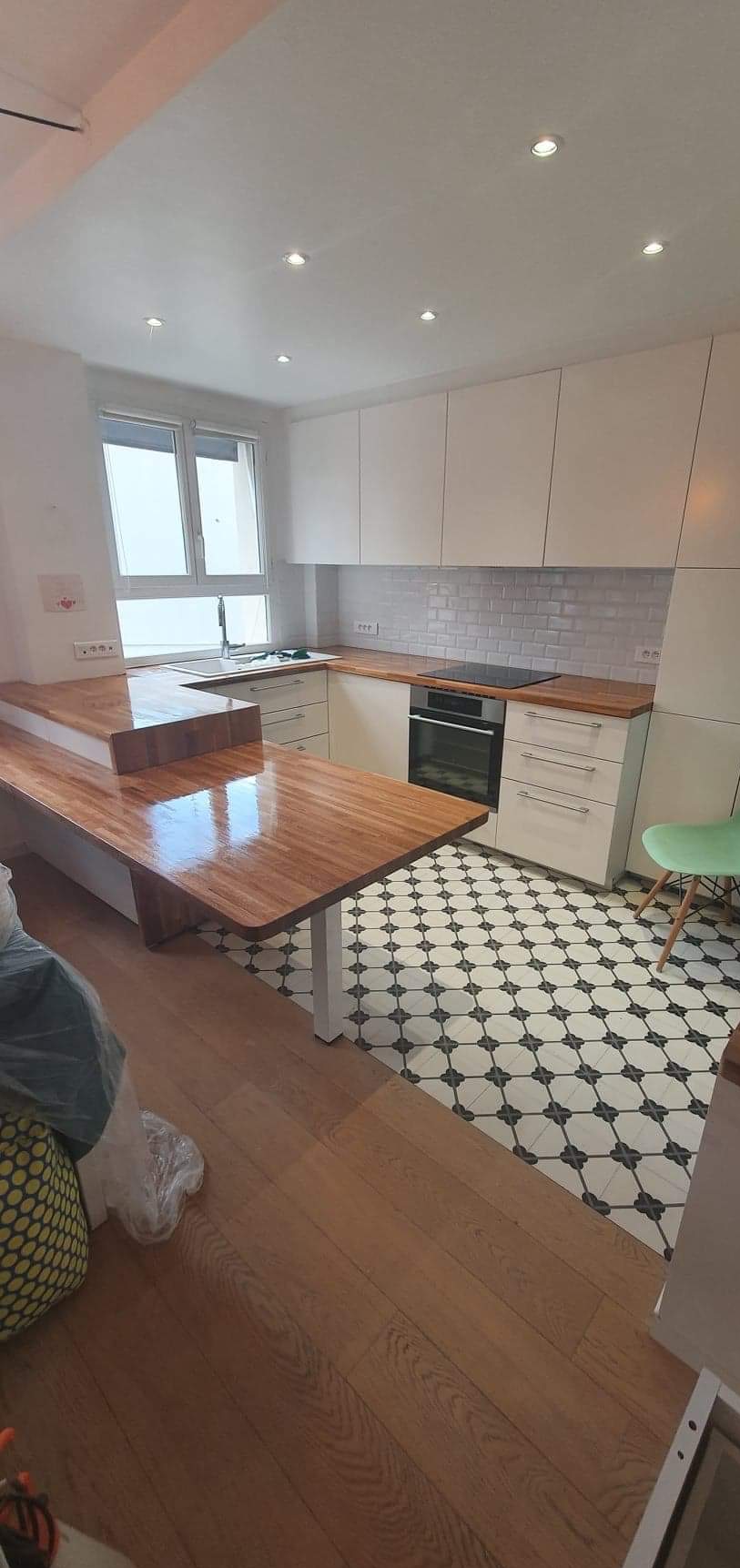 Nowoczesna kuchnia z drewnianym blatem i wzorzystą podłogą. Białe szafki, okno z widokiem na zewnątrz. Elementy wykończenia w stylu skandynawskim.