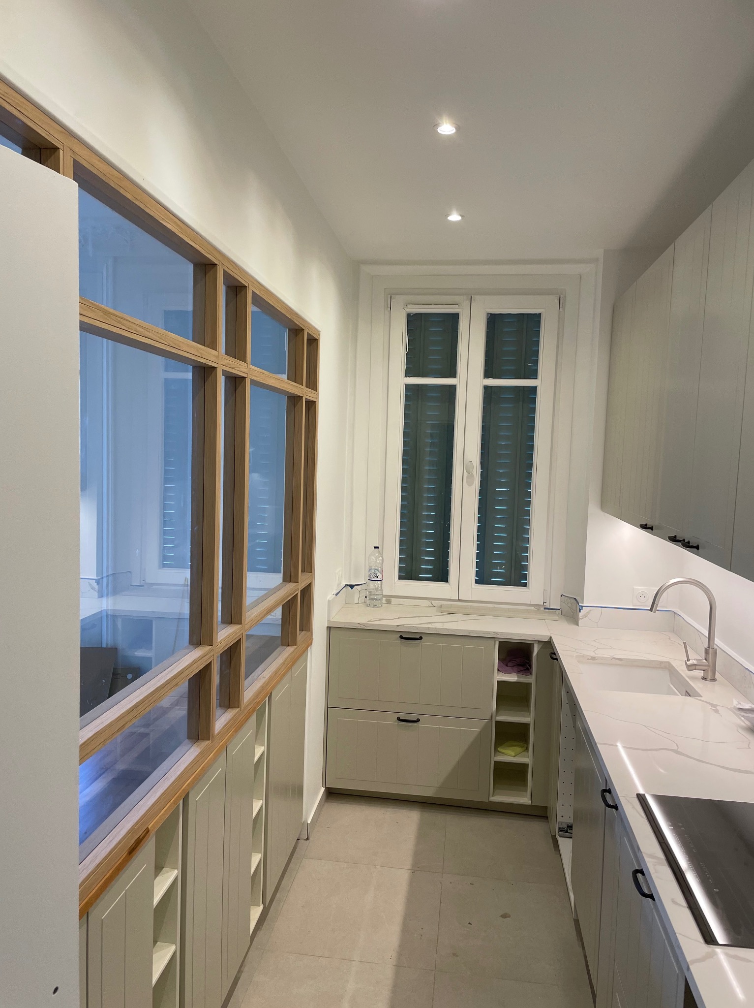 Wąska kuchnia w odcieniach szarości z drewnianą konstrukcją okienną nad szafkami. Blat z białego marmuru, nowoczesna armatura i oświetlenie punktowe.