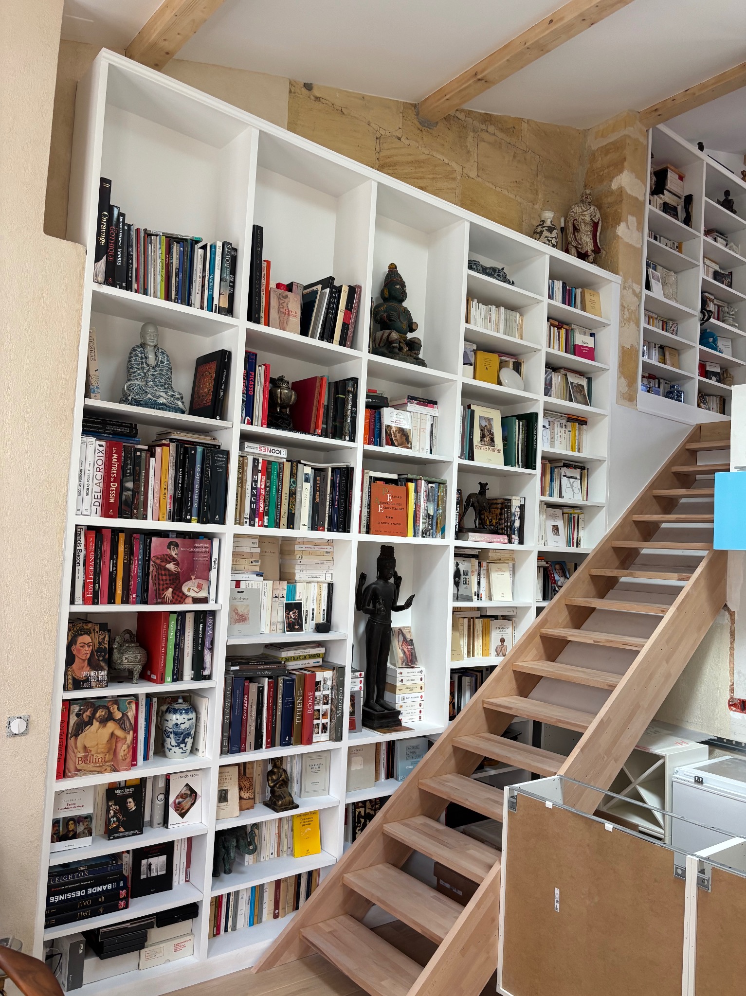 Biała, zabudowana biblioteka od podłogi do sufitu z drewnianymi schodami. Książki, dekoracje i naturalne światło tworzą przytulną atmosferę.