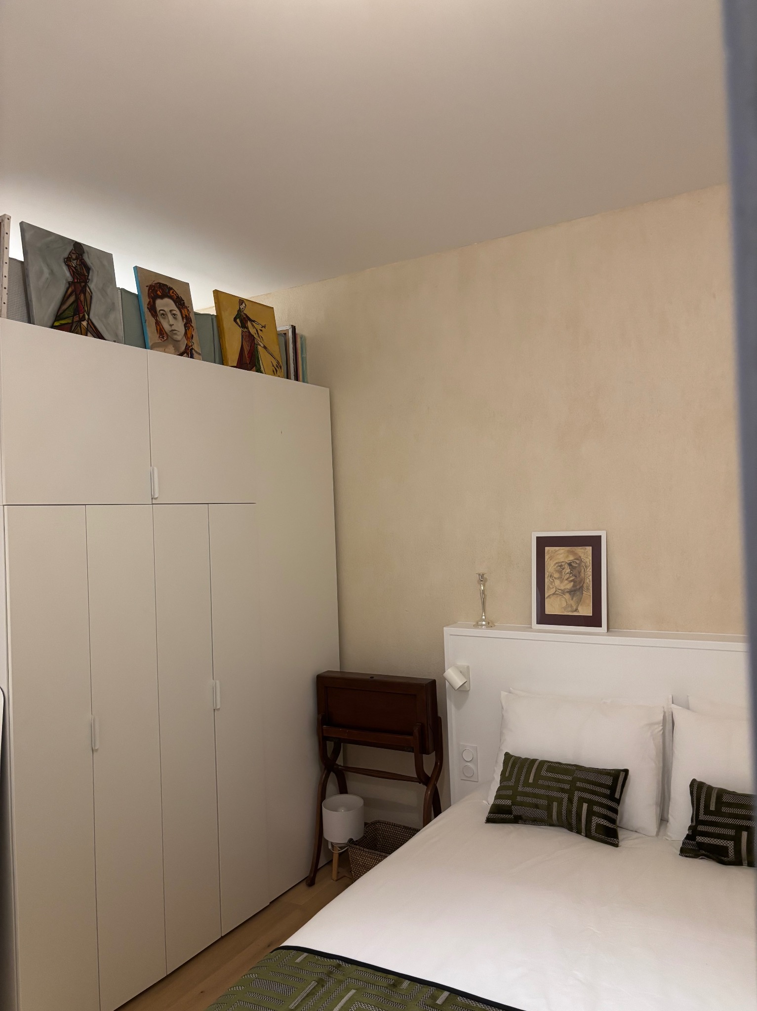 Jasne wnętrze sypialni z białymi meblami, obrazami na szafie i łóżkiem z wzorzystymi poduszkami. Minimalistyczny design z akcentami artystycznymi.