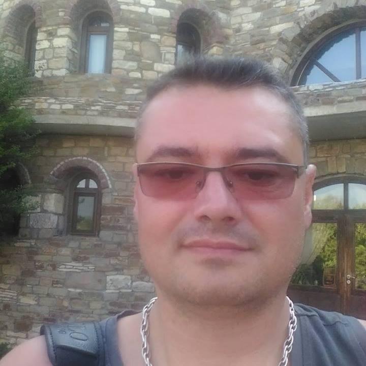 Mężczyzna w okularach przeciwsłonecznych i srebrnym łańcuszku na tle kamiennej budowli z łukowymi oknami.