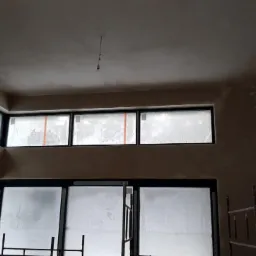 Wnętrze w trakcie prac wykończeniowych: ściany po nałożeniu świeżego tynku, okna zabezpieczone folią, widoczna instalacja elektryczna i fragment rusztowania.