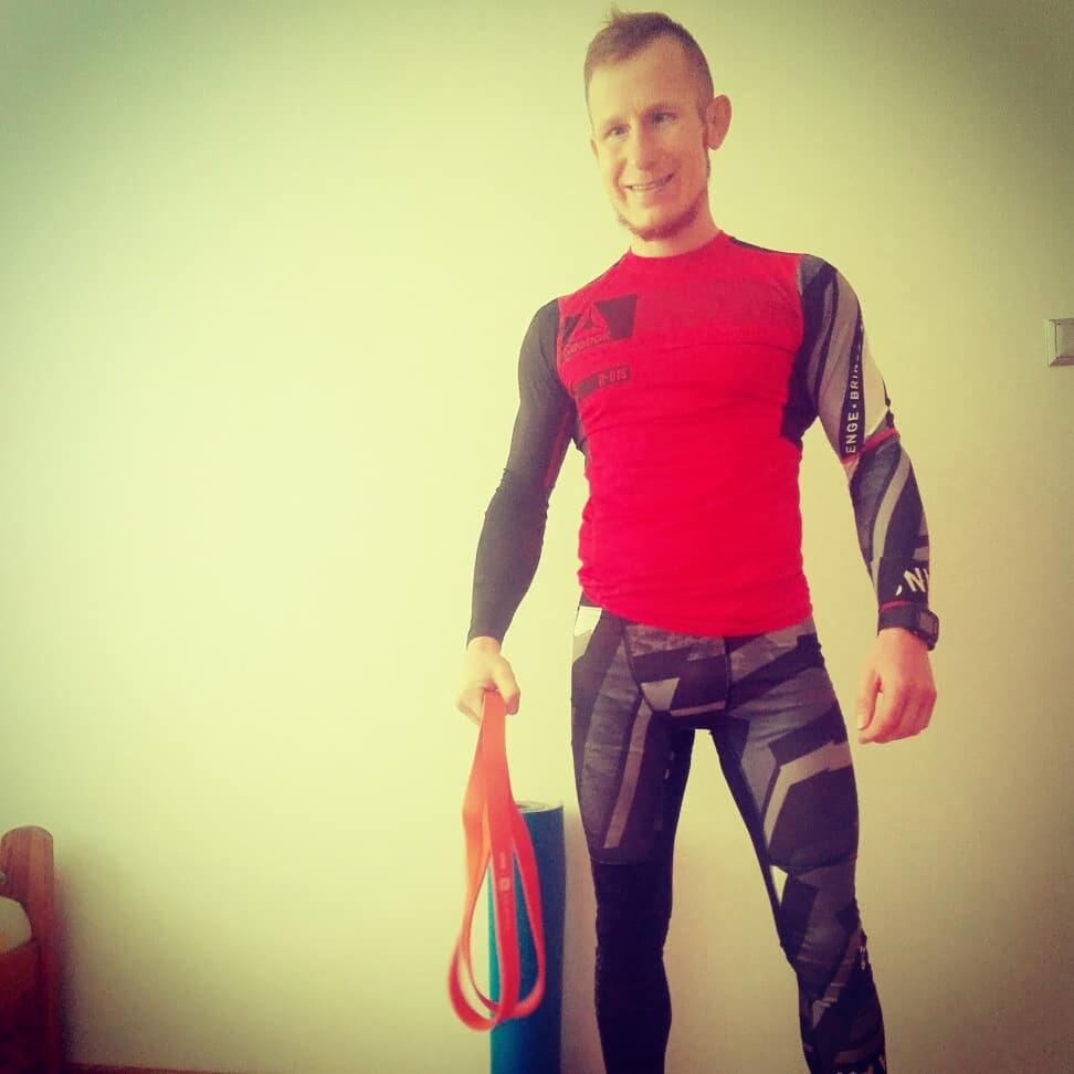 Uśmiechnięty mężczyzna w sportowym stroju trzyma gumę do ćwiczeń, widoczna mata do ćwiczeń w tle.