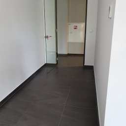 Łukasz Grzegrzółka - Wnętrze biura z otwartymi szklanymi drzwiami, ciemną podłogą z płytek i białą ścianą z hydrantem.