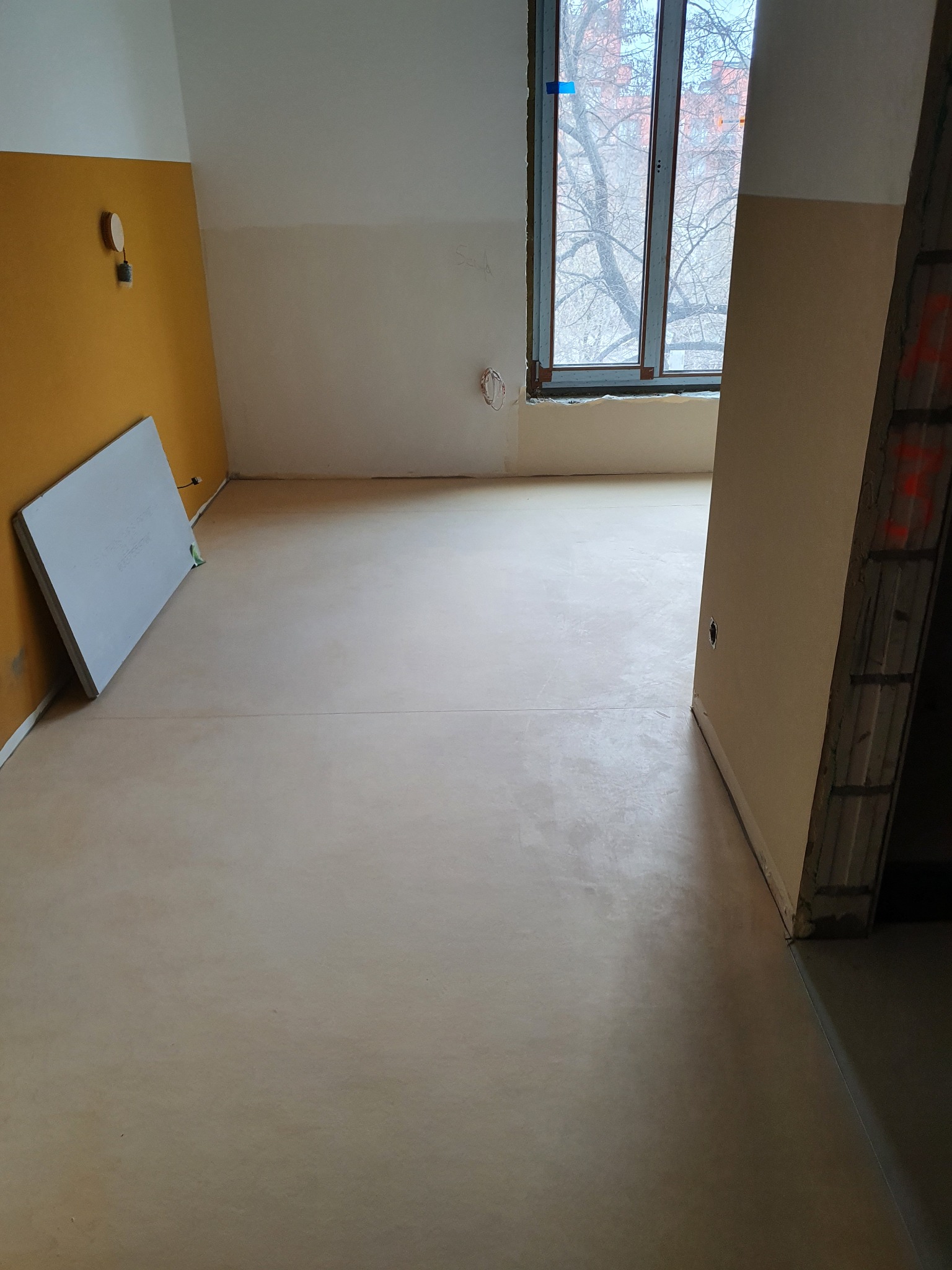 Wnętrze po wylewce samopoziomującej, widoczne ślady po remoncie, okno z widokiem na drzewo bez liści.