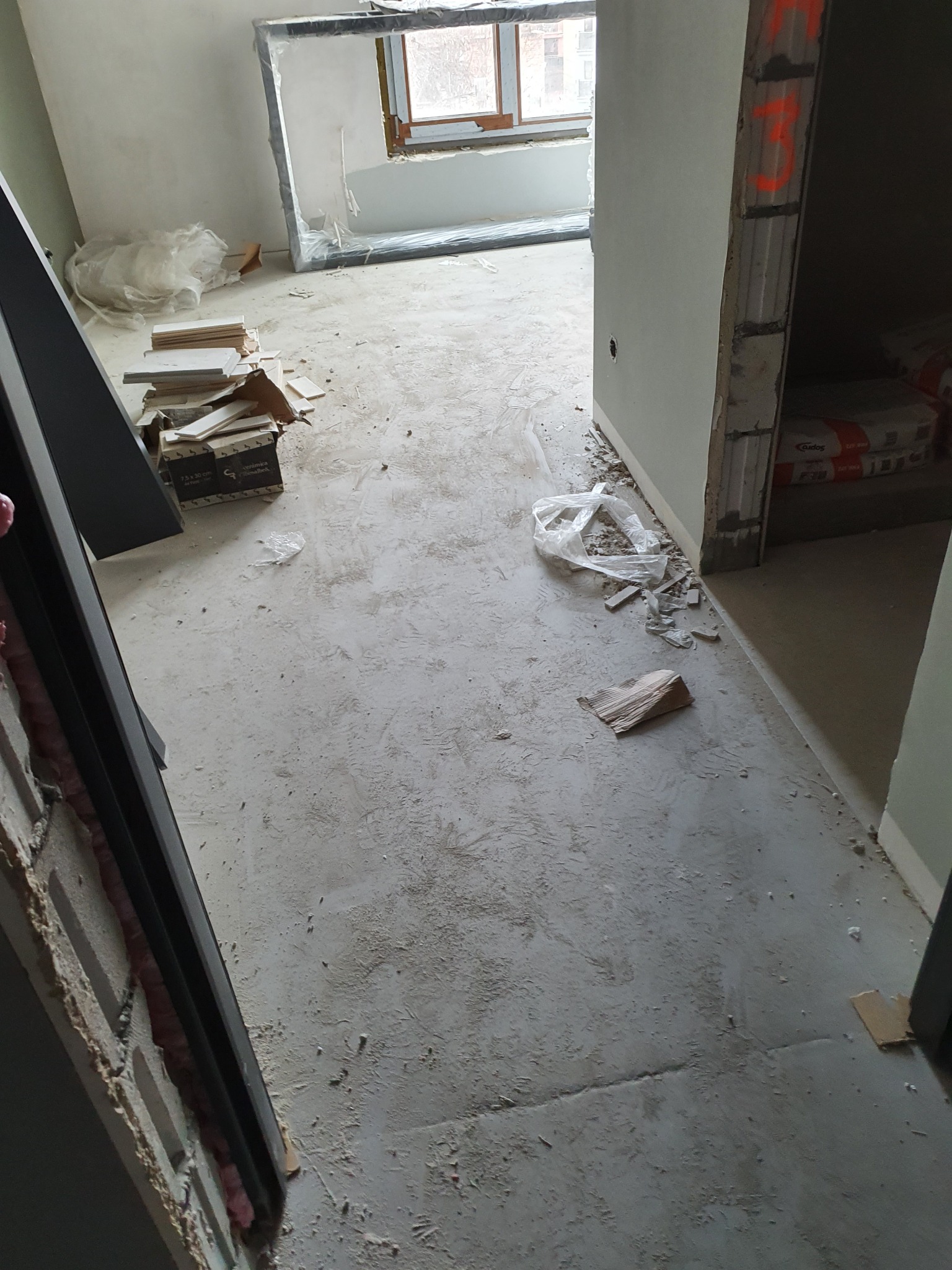 Wnętrze w trakcie remontu: rozsypane płytki, gruz na podłodze, widoczne worki z zaprawą, okno z widokiem na budynki.