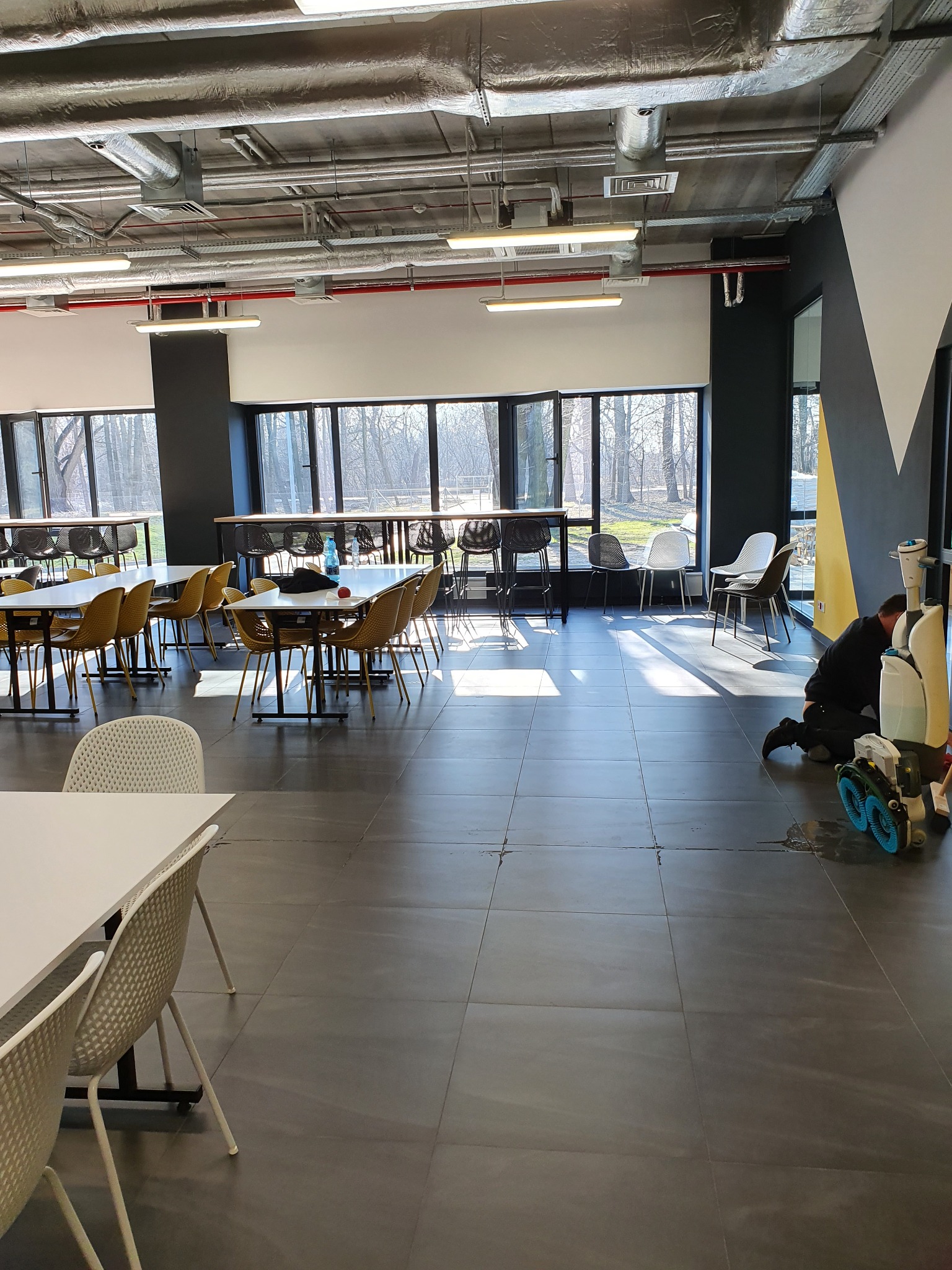 Pracownik w czarnym ubraniu klęczy na podłodze, obsługując maszynę do mycia podłóg z niebieskimi szczotkami w przestronnej stołówce biurowej z rzędami stołów i krzeseł oraz dużymi oknami.