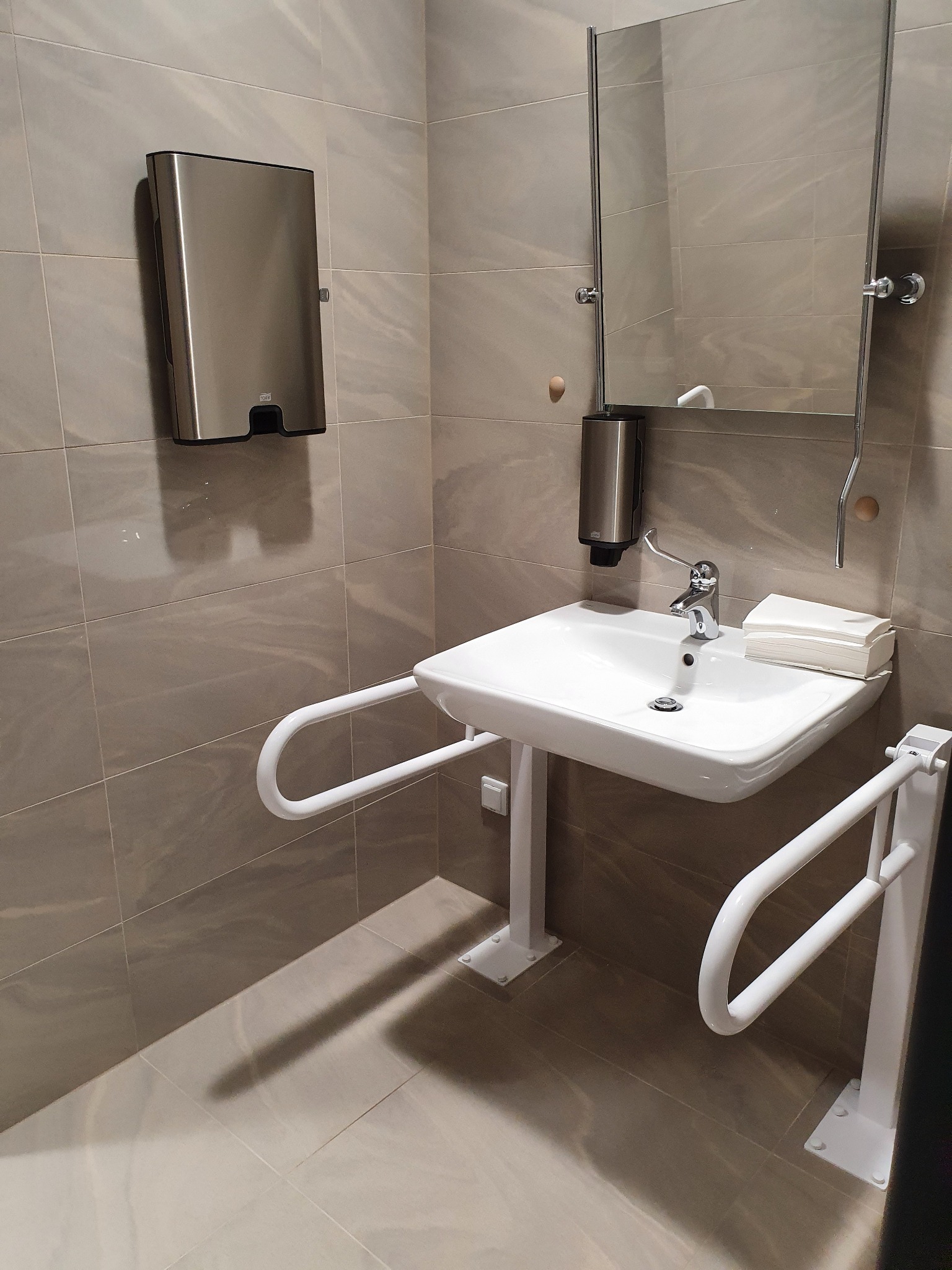 Toaleta dla niepełnosprawnych z umywalką, uchwytami, dozownikiem mydła i papieru oraz lustrem, wykończona w szaro-beżowych płytkach.