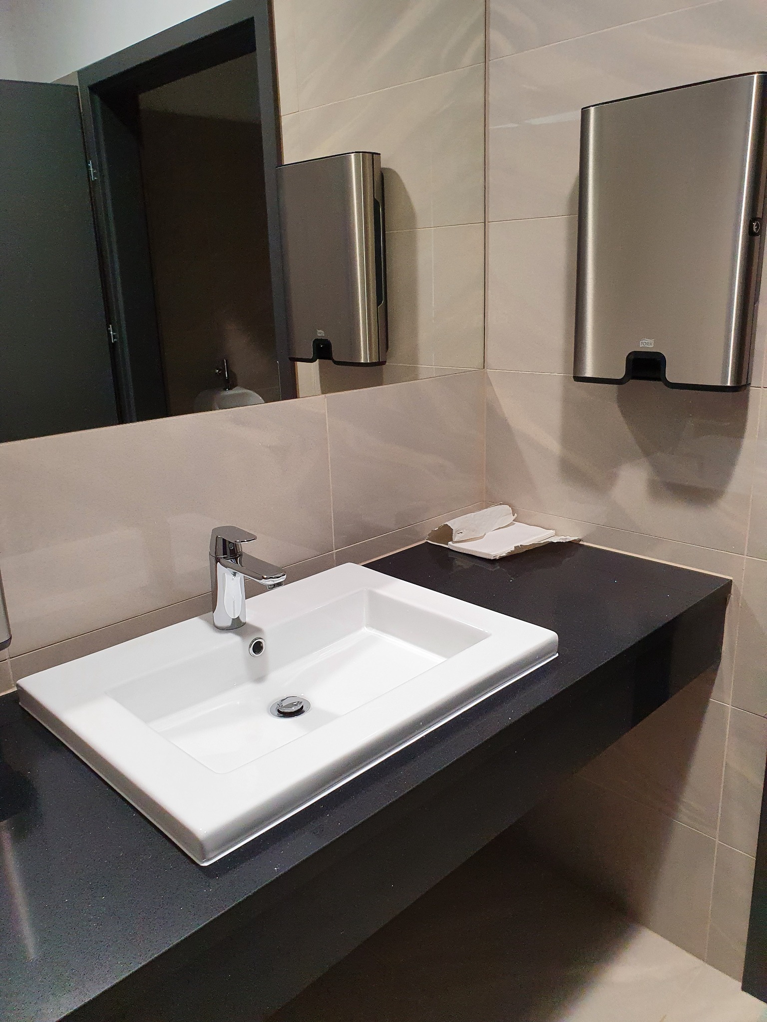 Biała umywalka z chromowaną baterią na czarnym blacie w toalecie biurowej, z widocznym dozownikiem ręczników papierowych i lustrem.
