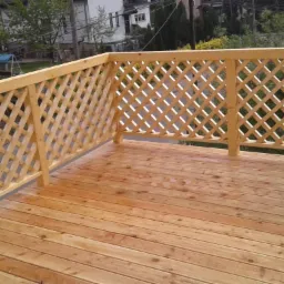 Nowo wybudowany, drewniany taras z kratownicową balustradą, ocieplony odcieniami brązu, z widokiem na zielony ogród i zabudowę jednorodzinną w tle.