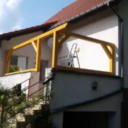 Żółta drewniana konstrukcja zadaszenia balkonu w trakcie budowy, widoczna drabina oparta o ścianę budynku z czerwoną dachówką, częściowo zasłonięta przez bujną zieleń.