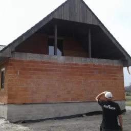 Budowa domu z czerwonej cegły w trakcie realizacji, widoczny fundament z szarej cegły, niezabezpieczone okna oraz mężczyzna w koszulce z logo firmy budowlanej obserwujący postęp prac.