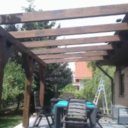 Drewniana pergola z belek, zacieniająca taras z meblami ogrodowymi, widok na dom z cegły i ogród.
