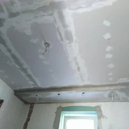 Niedokończony sufit z widocznymi łączeniami płyt gipsowo-kartonowych przed szpachlowaniem, wystające przewody elektryczne, surowe ściany z widocznymi instalacjami elektrycznymi i oknem.