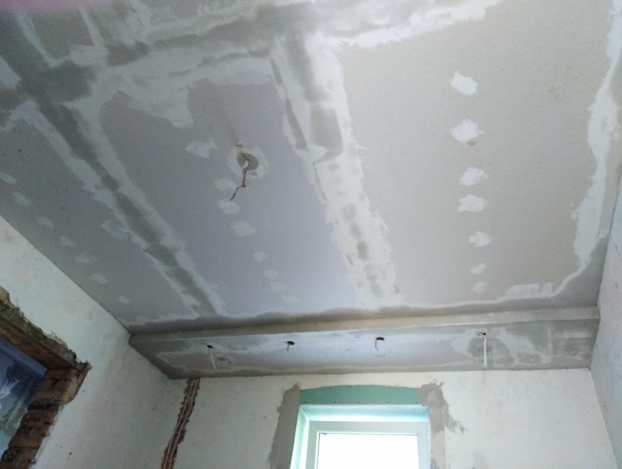 Niedokończony sufit z widocznymi łączeniami płyt gipsowo-kartonowych przed szpachlowaniem, wystające przewody elektryczne, surowe ściany z widocznymi instalacjami elektrycznymi i oknem.