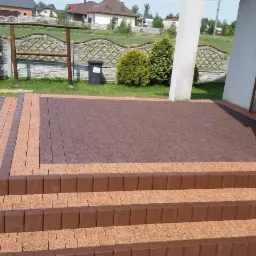 Ułożona kostka brukowa w dwóch odcieniach brązu tworząca schody i taras przed domem, widoczne elementy ogrodzenia i zieleń w tle.