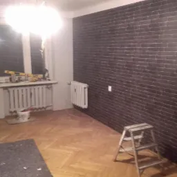 Ściana wykończona tapetą imitującą cegłę, z widoczną drabiną malarską, poziomica na parapecie i wiadro z narzędziami w rogu pokoju.