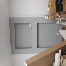 Bonabud - Wykończona ściana z dwoma wnękami pomalowanymi na szaro, w tle kartony, drabina i narzędzia malarskie, widoczne przewody elektryczne.