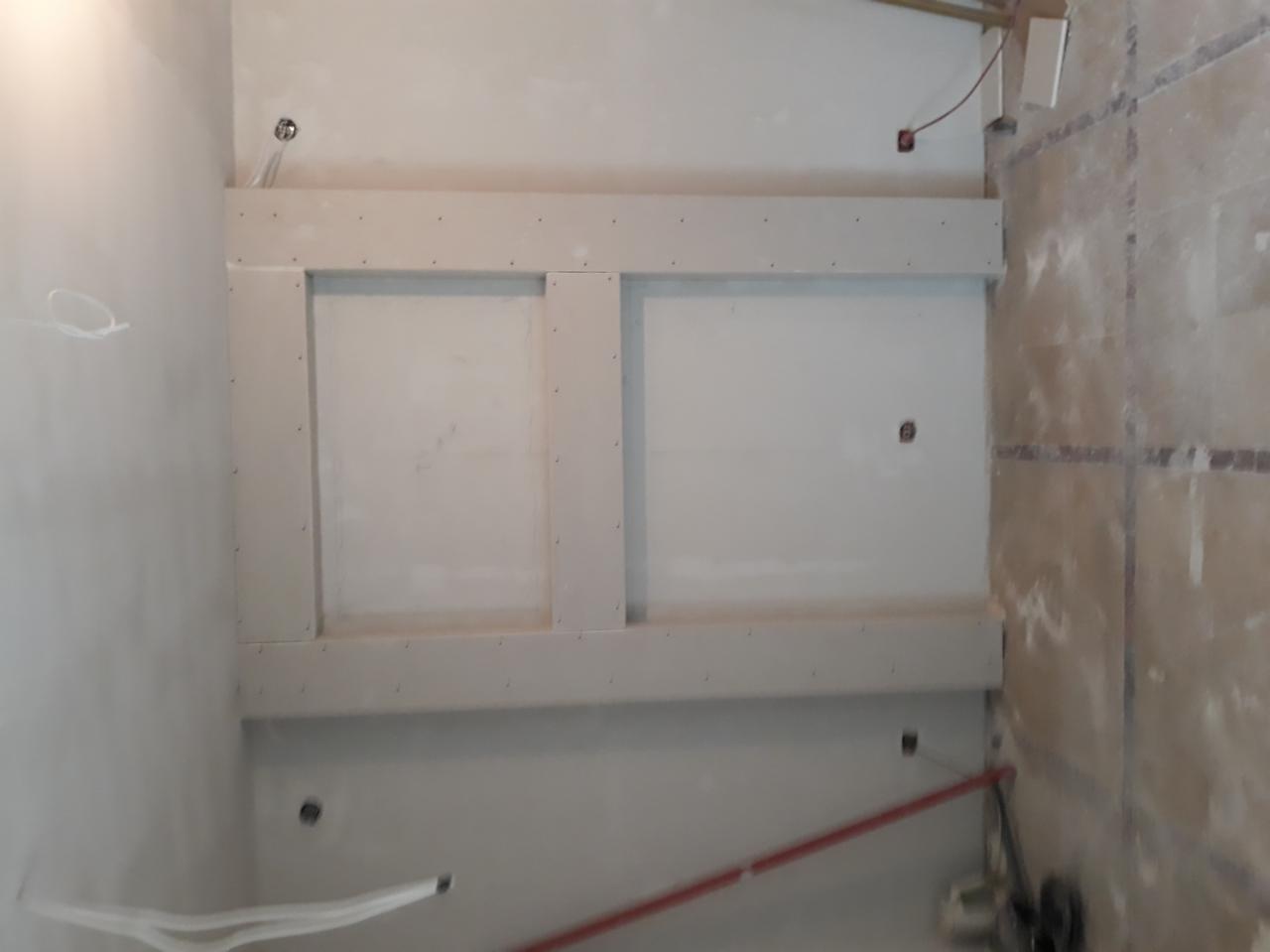 Konstrukcja z profili i płyt na ścianie, przygotowana pod zabudowę, widoczne otwory na instalacje, fragment ściany wykończonej płytkami w tle.