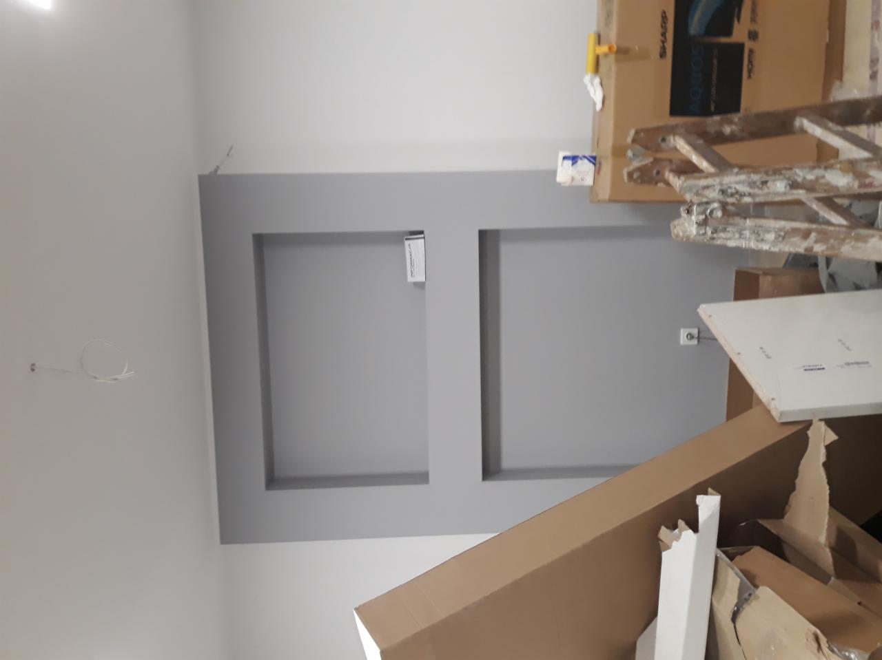 Wykończona ściana z dwoma wnękami pomalowanymi na szaro, w tle kartony, drabina i narzędzia malarskie, widoczne przewody elektryczne.