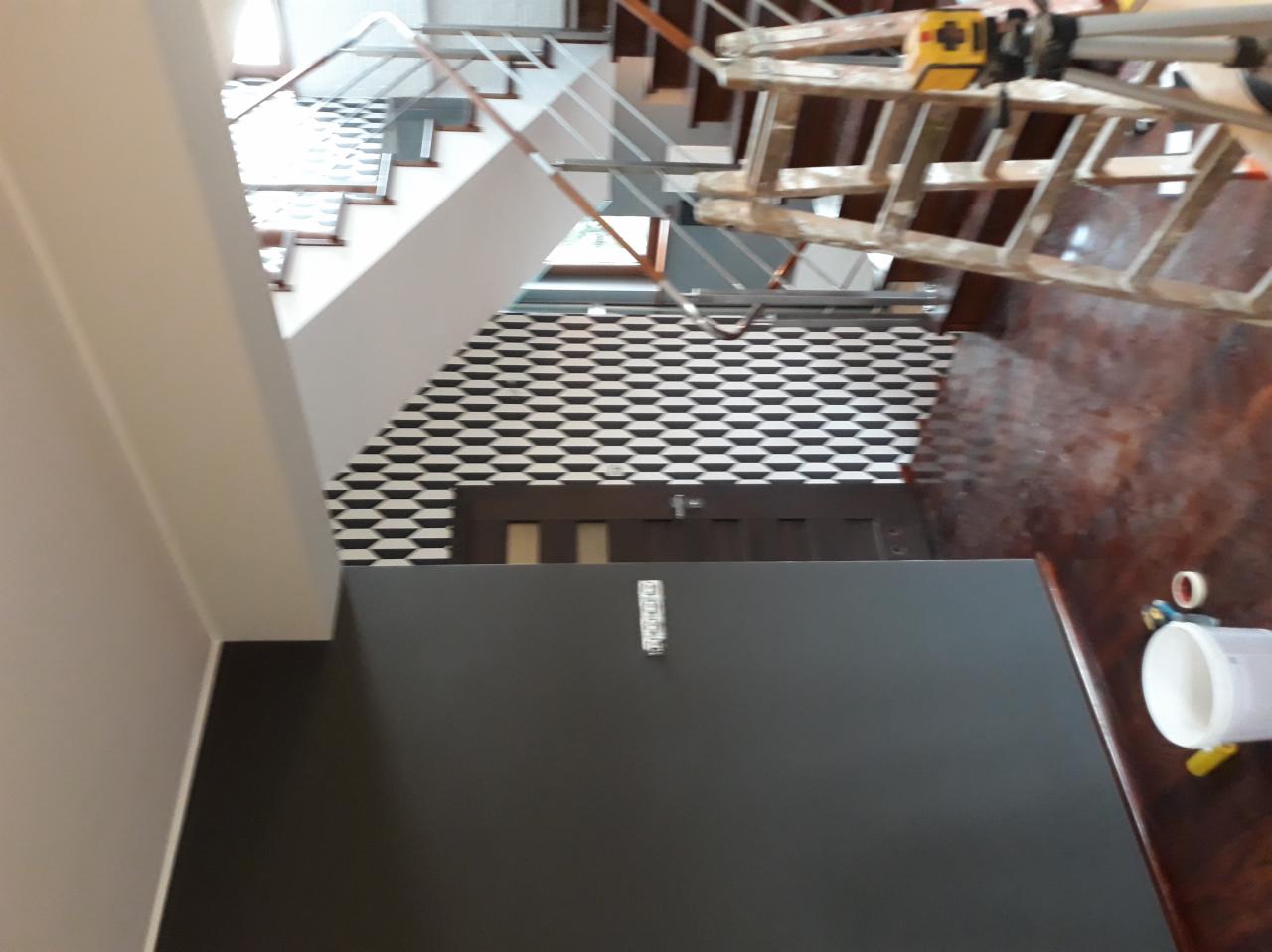 Ściana pomalowana na ciemny kolor w korytarzu ze schodami, widoczna drabina malarska i puszka farby, ściana z wzorem geometrycznym w tle.