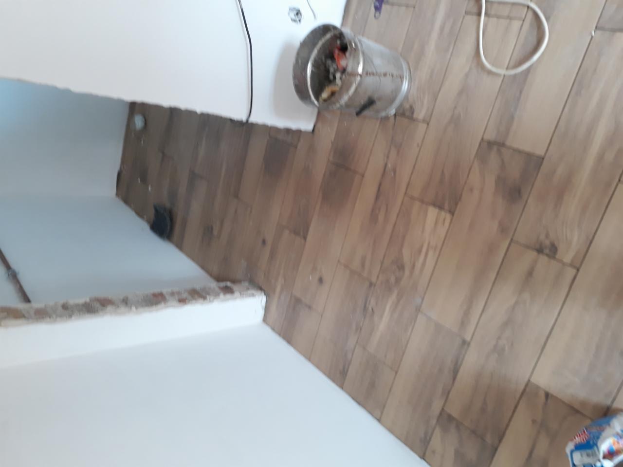 Fragment pomieszczenia w trakcie remontu: świeżo pomalowana ściana z widocznymi śladami po wcześniejszych pracach, podłoga wyłożona panelami imitującymi drewno, metalowe wiadro z odpadami budowlanymi.