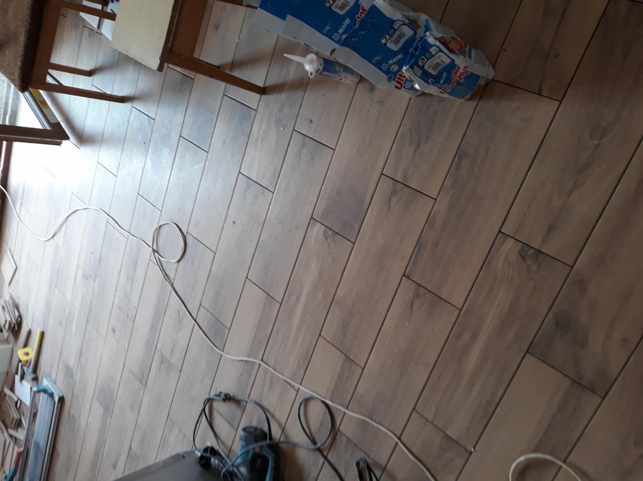 Ułożona podłoga z płytek imitujących drewno, widoczne narzędzia glazurnicze, klej i fuga w opakowaniach, krzesło w rogu.